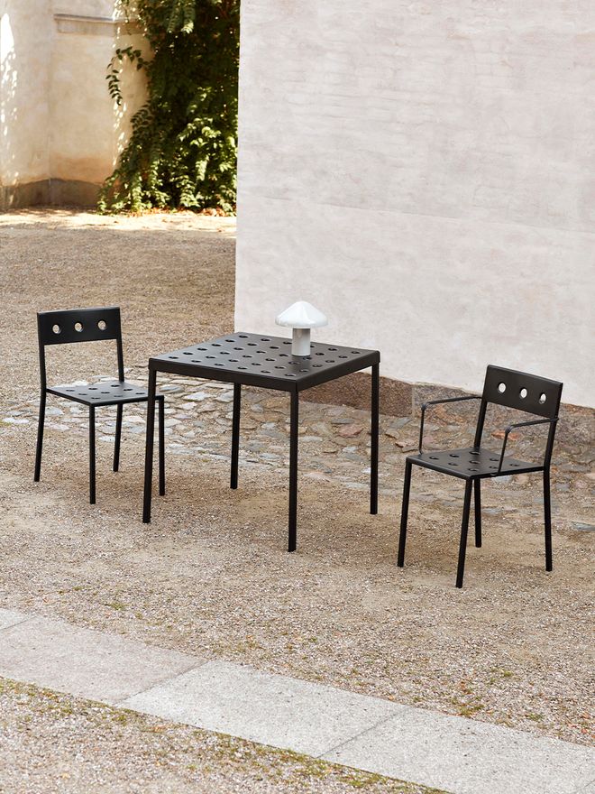 HAY’s Balcony table and two chairs in a courtyard next to a building. There is also a white, mushroom-shaped Pao Portable table lamp on the table.
