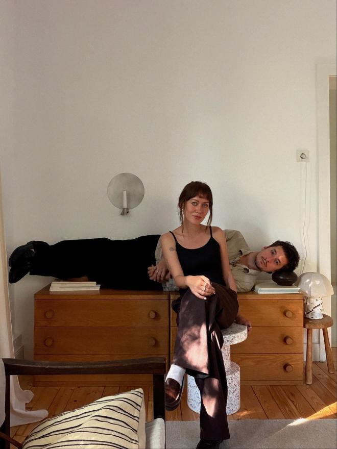 Isabelle Kaiser sits in front of a wooden dresser while Bari Chyba lies across the top. A pale wall and a small round wall lamp are behind them.