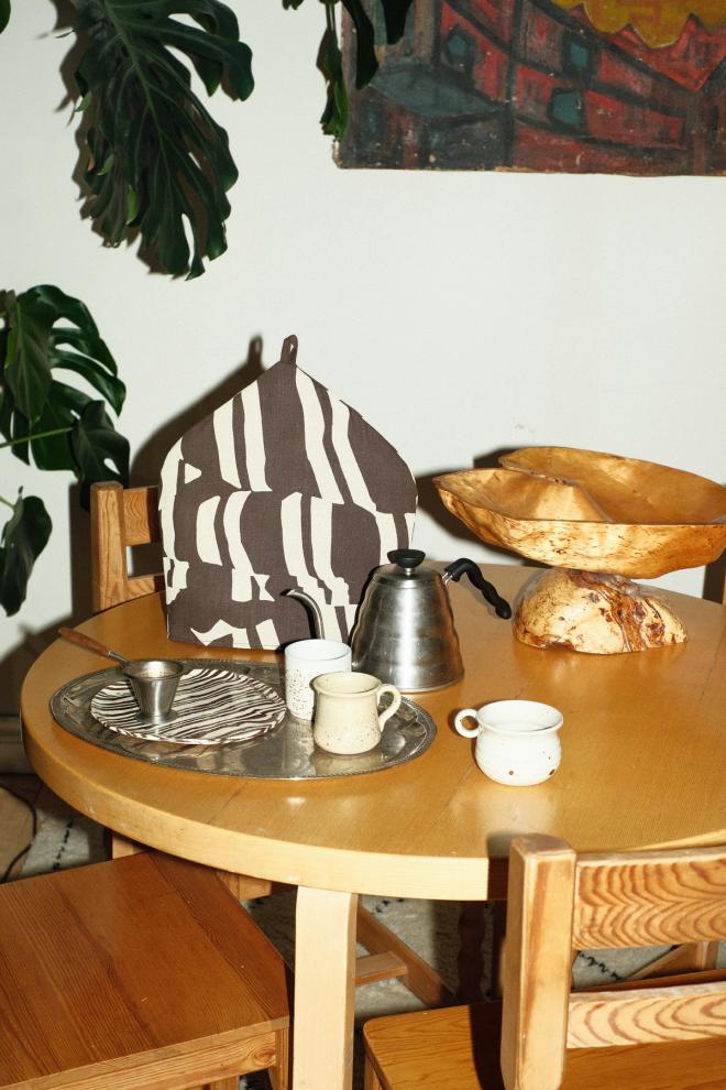 A round wooden table set with a teapot covered by a black-and-white patterned tea cosy, light-coloured cups, bread in a wooden bowl, and a houseplant in the background