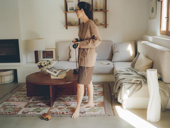 A woman stands in a bright living room holding a camera, looking down at it. A low wooden table, a pale sofa, wooden beams across the ceiling, and a children's play mat with a toy car on the floor.