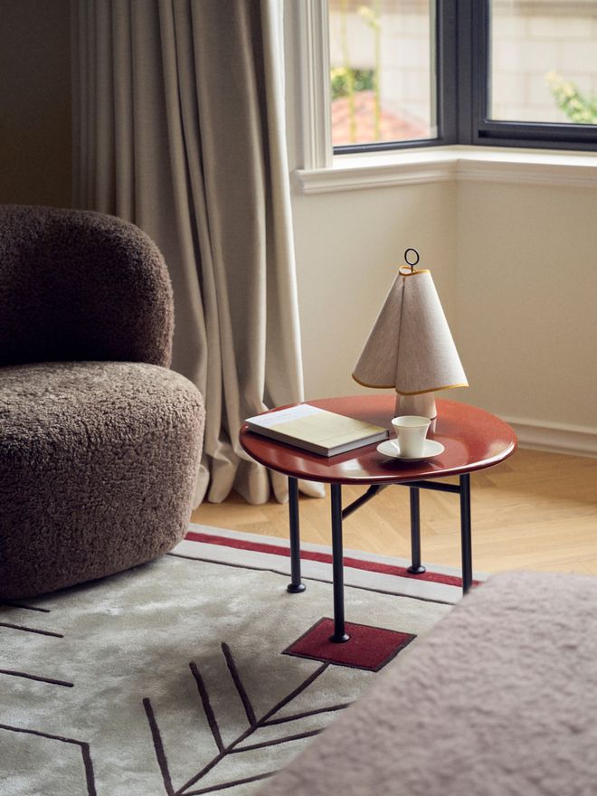 Round red side table with black legs holding a fabric-shaded table lamp, a book, and a teacup, beside a textured armchair near a curtained window.