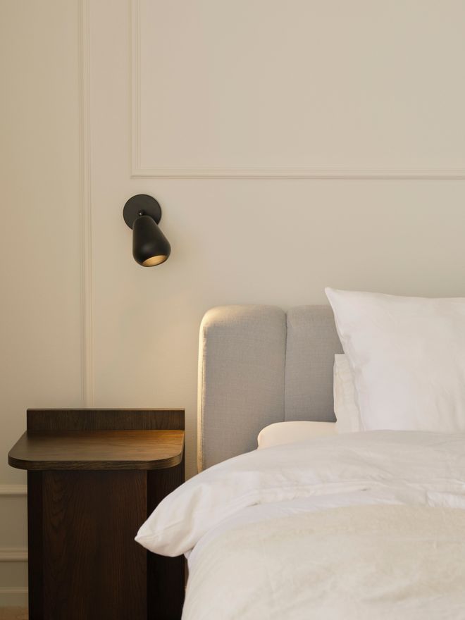 A close-up of the head of a bed: a pale upholstered headboard, white linens, a dark wooden bedside table with a small black spotlight on the wall.