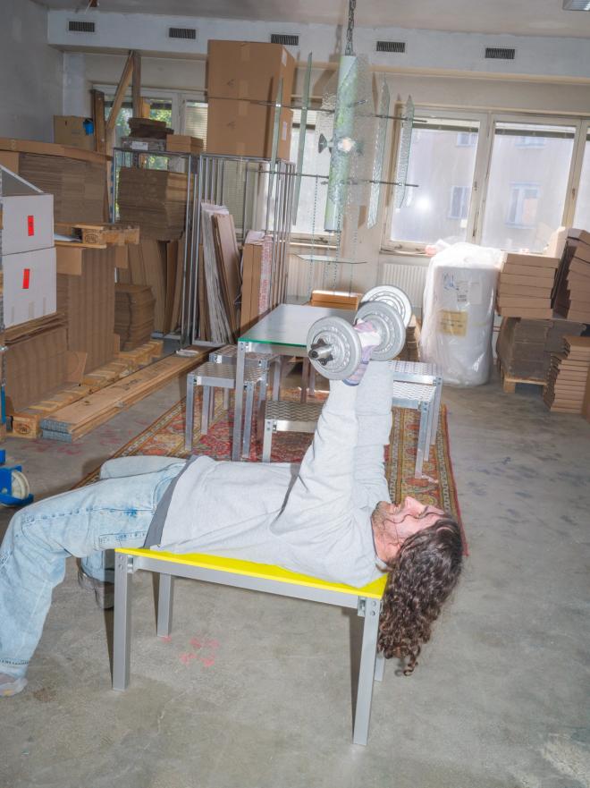 Fredrik Paulsen lies on his back on a yellow bench lifting dumbbells in a workshop, surrounded by timber and cardboard boxes.