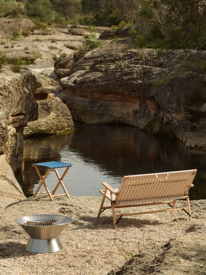 A mountain landscape featuring a sun lounger, table, and outdoor fire pit from HAY’s Outdoor Market collection. Water can be seen in the background. 