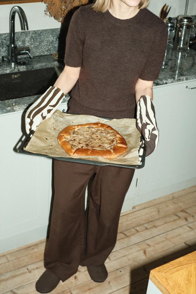 A person holding a baking tray with a freshly baked pie, wearing black-and-white patterned oven mitts, with a kitchen sink in the background