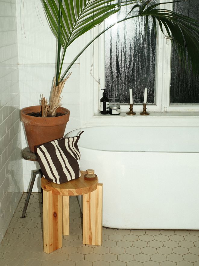 A black-and-white striped toiletry bag on a wooden stool beside a bathtub, with a terracotta planter, candleholders, and a window in the background.