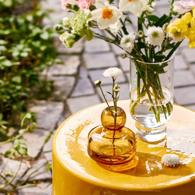 Marimekko's amber colored Flower vase and Karakter's Cessidra vase filled with flowers, standing on Serax's ochre colored Pawn side table.