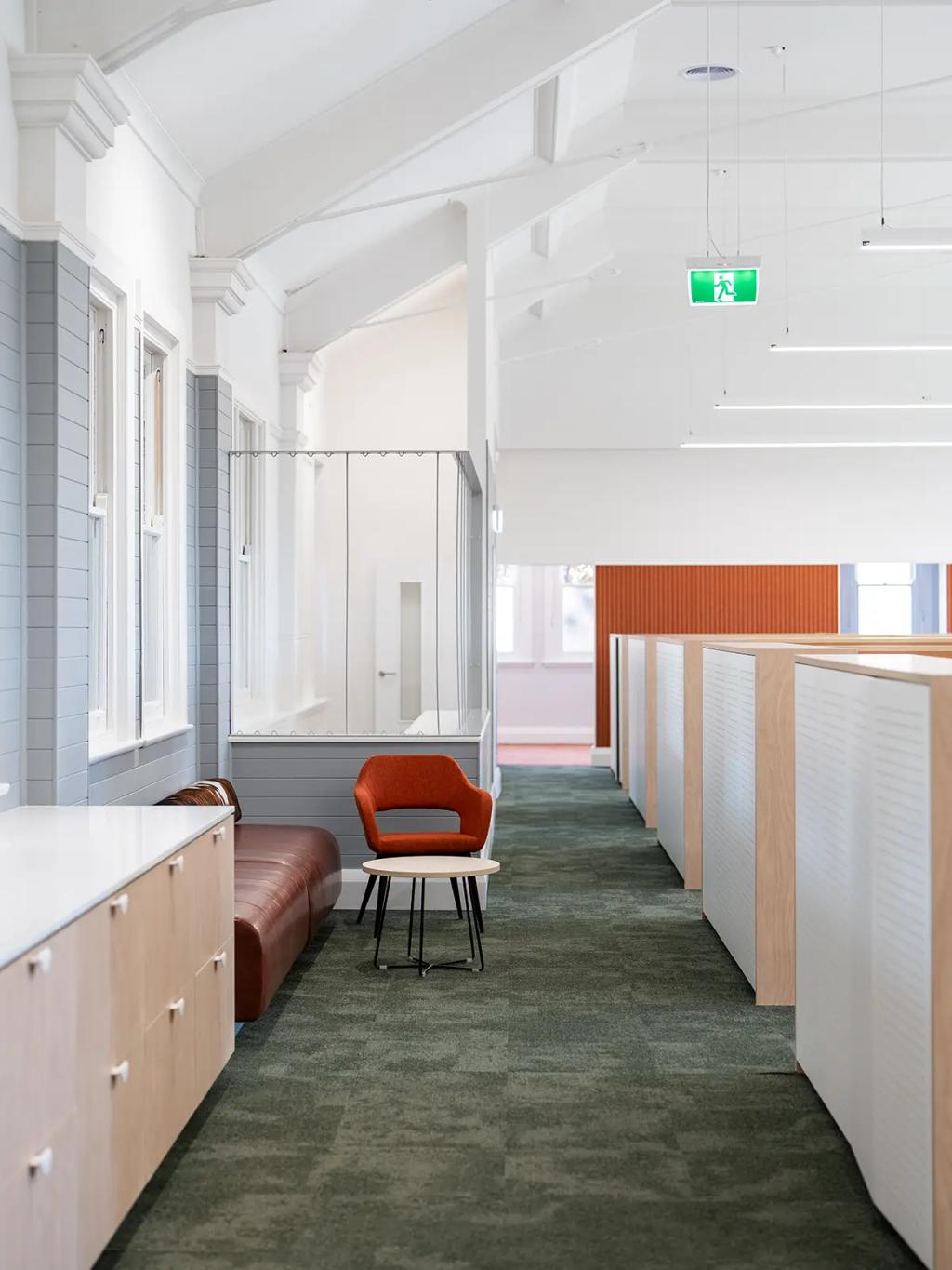 Downlands College Toowoomba Refurbishment (Hallway lounge area with a brown leather bench, orange accent chair, round table, and overhead exit sign.)