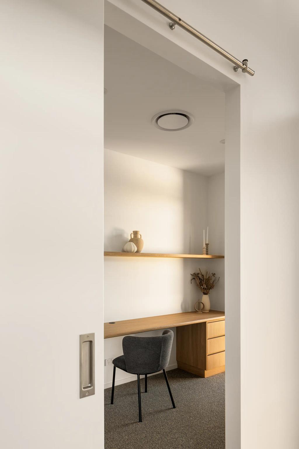 Project Ward (A home office with a wooden desk, grey chair, floating shelves, and minimalist decor, seen through a doorway)