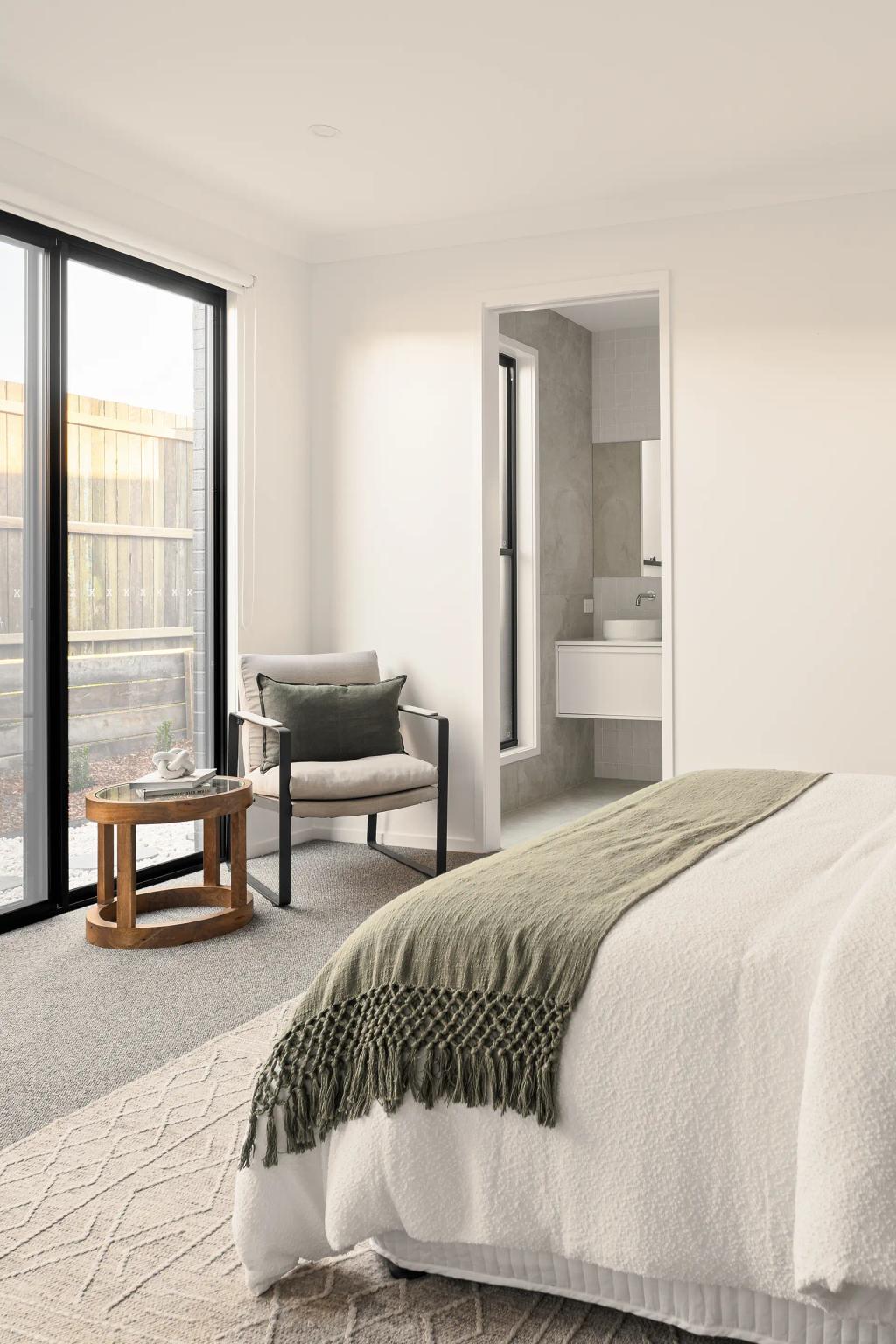 Project Ward (Bright bedroom with a cozy chair, wooden side table, and bed with a green throw, leading to a bathroom)