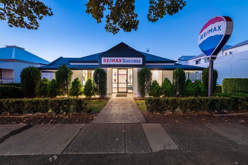 Project RE/MAX (RE/MAX office exterior at dusk with glass entrance, hedges and hot-air-balloon pylon sign)
