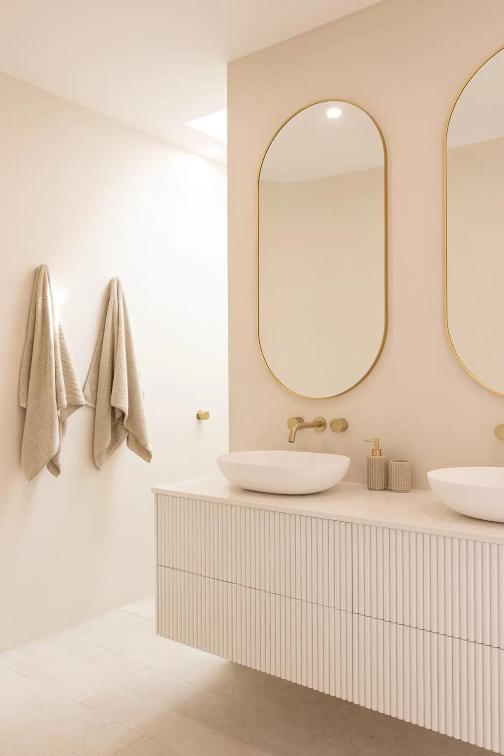 Project Vuse (A modern bathroom with beige walls, featuring two oval-shaped mirrors with gold frames above a white vanity with ribbed drawers)