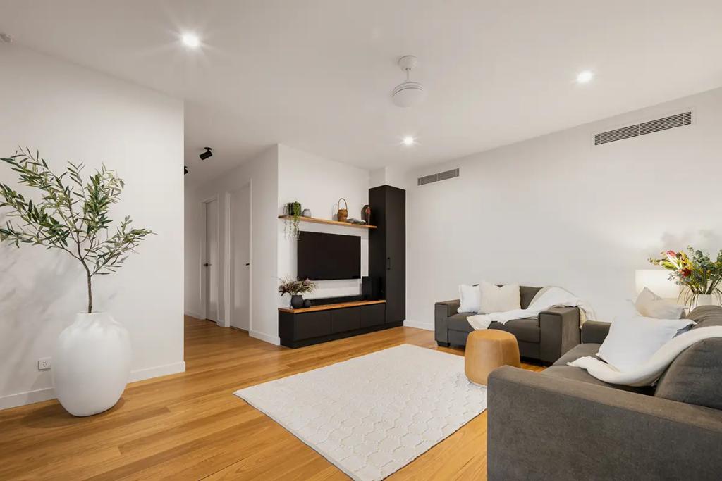 Project Treevue (Modern living room with white walls, wooden floors, a large olive tree in a white vase, a dark gray sectional sofa with throw pillows, a white area rug, and a built-in black entertainment unit with a flat-screen TV)