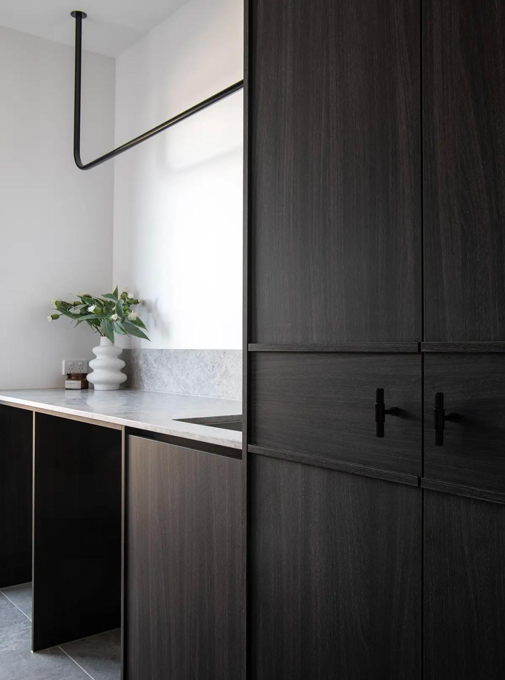 Project Ward 2.0 (A modern laundry room with dark wood cabinetry, a marble countertop, a black hanging rod, and a white vase with greenery)