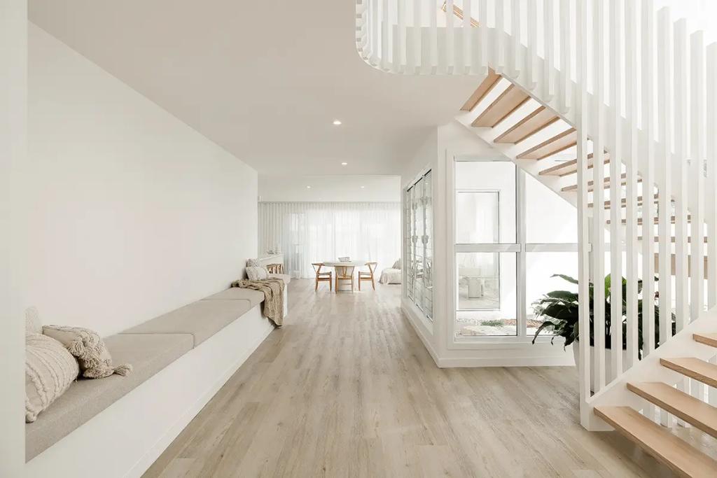 Project Vuse (A modern, minimalist Interior with light wood floors, white walls, a built-in bench with cushions, a wooden dining table with chairs, and a white staircase with vertical slats)