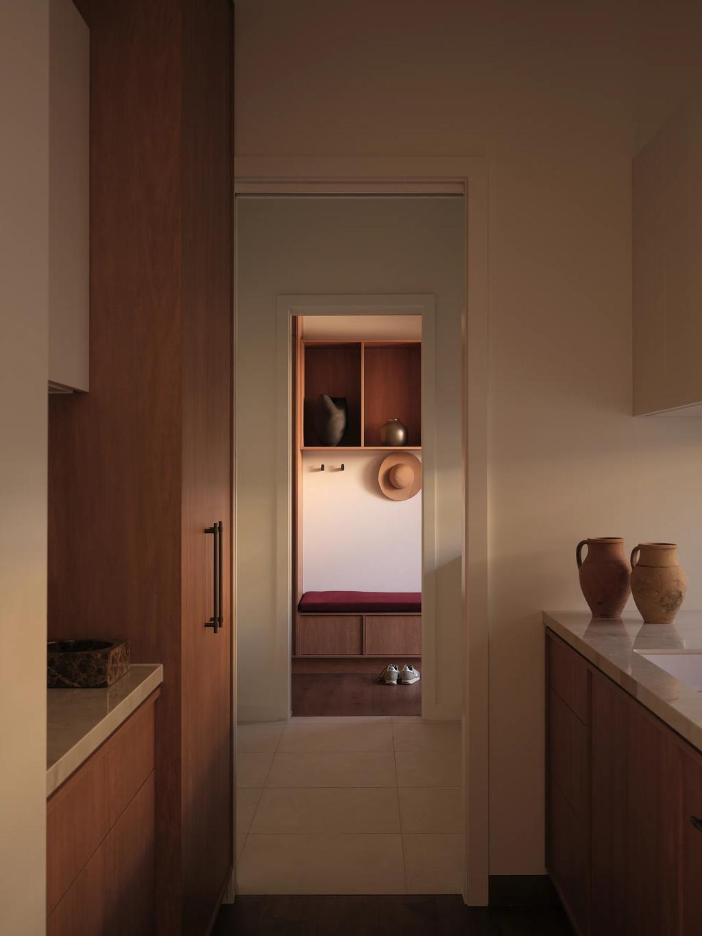 Project Riverton (Hallway view into a mudroom with a straw hat, shoes, and a bench, flanked by kitchen counters.)