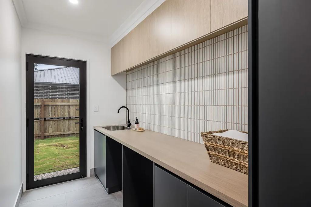 Modern kitchen with light wood cabinetry, black countertops and black sink faucet, white tiled backsplash, and a glass door leading to the backyard