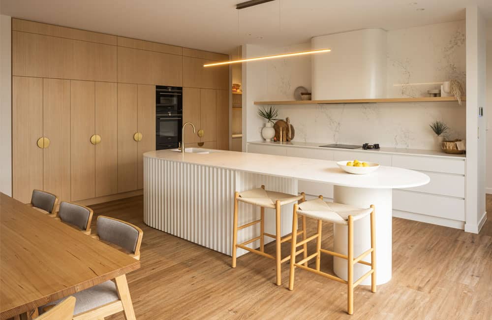 San Clemente (White fluted island, light timber cabinetry with gold pulls and linear pendant light)
