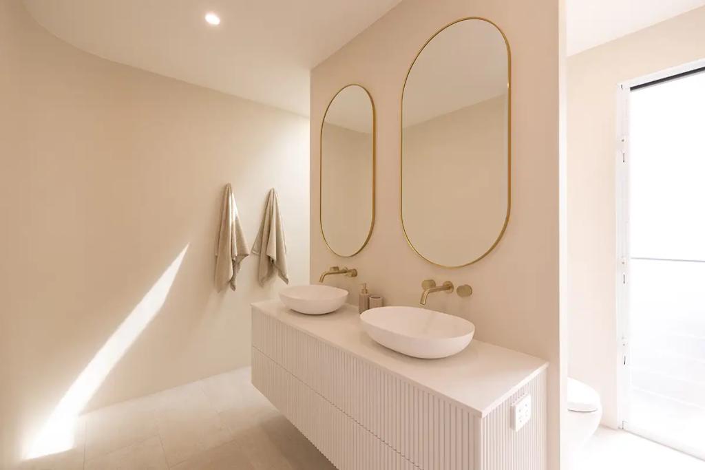 Project Vuse (Modern minimalist bathroom with two oval mirrors, white bowl sinks on a wooden vanity, and beige towels hanging on the wall)