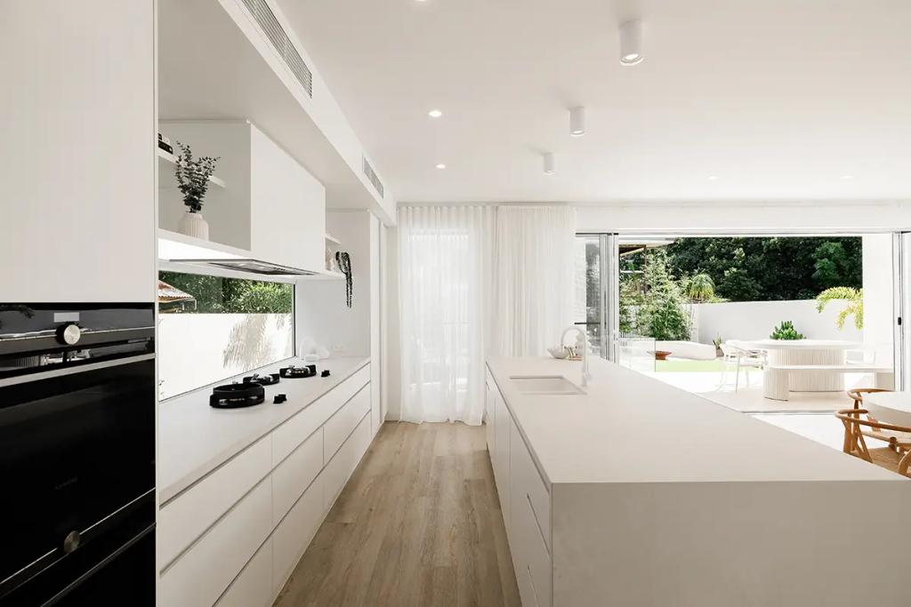 Project Vuse (Modern kitchen with white cabinets, countertops and appliances, light wood flooring, and a view of an outdoor patio through sliding glass doors with sheer curtains)