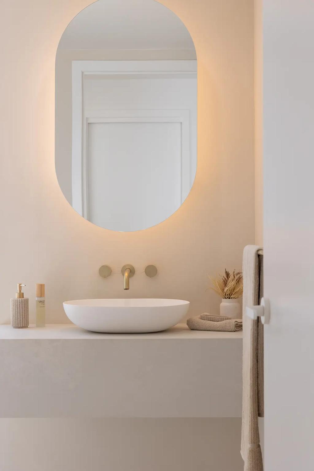 Project Vuse (A minimalist bathroom with a round mirror, white vessel sink with golden faucet, and beige countertop with soap dispenser, small vase with dried plants, and folded towels)