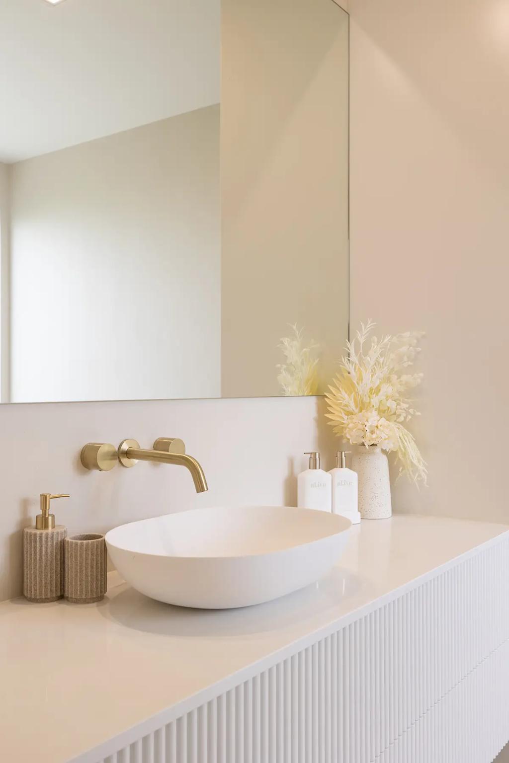 Project Vuse (A modern bathroom with a white oval-shaped sink with a gold faucet and a vase with white and yellow dried flowers)
