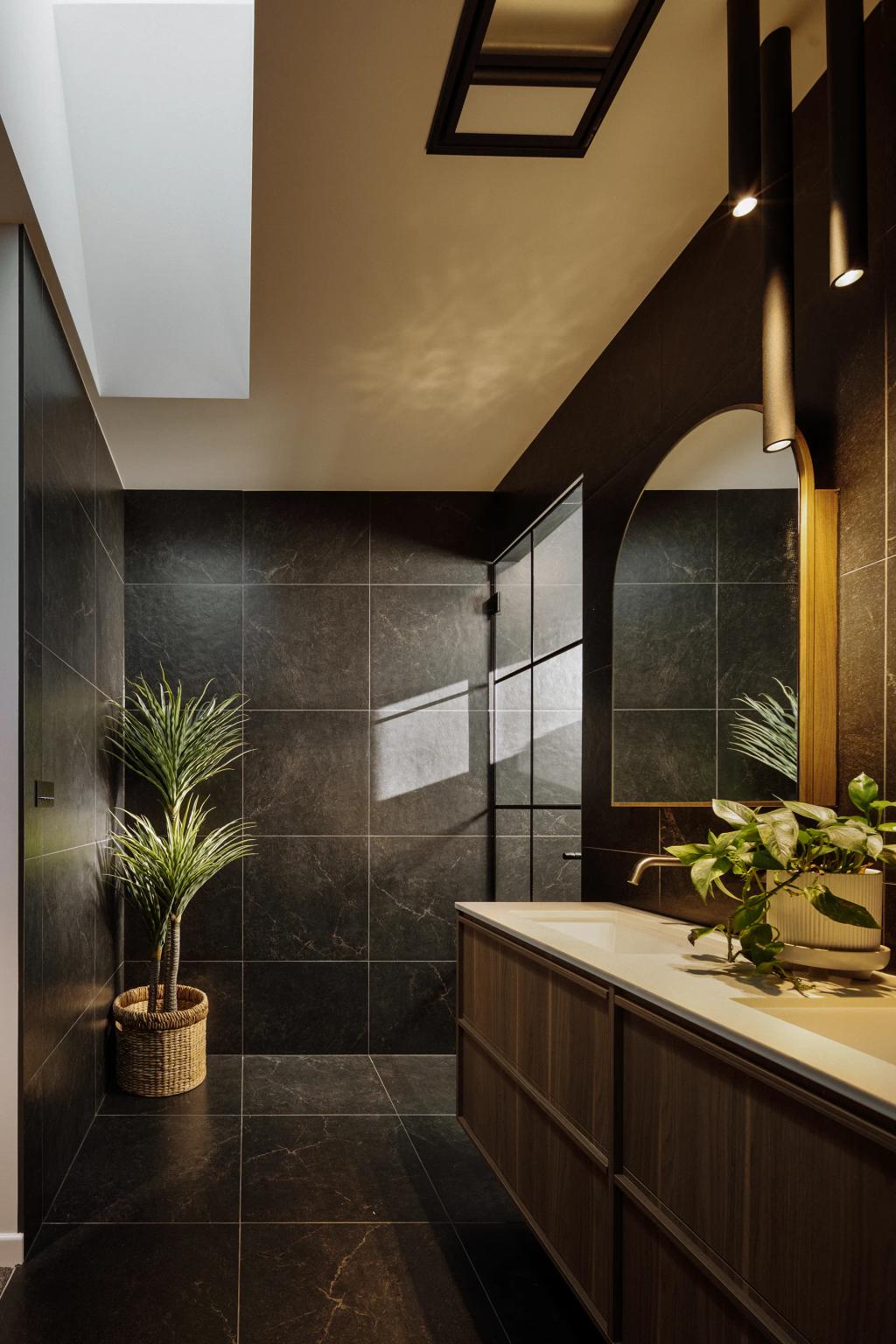 Lavish Morada (Elegant bathroom with black tile walls, wood vanity, arched mirror, and natural lighting.)