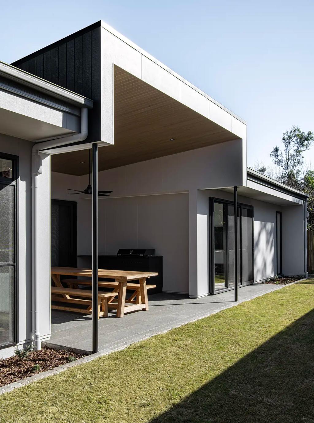 Project Ward 2.0 (A modern outdoor patio with a wooden picnic table, barbecue grill, covered ceiling, and neatly trimmed lawn)