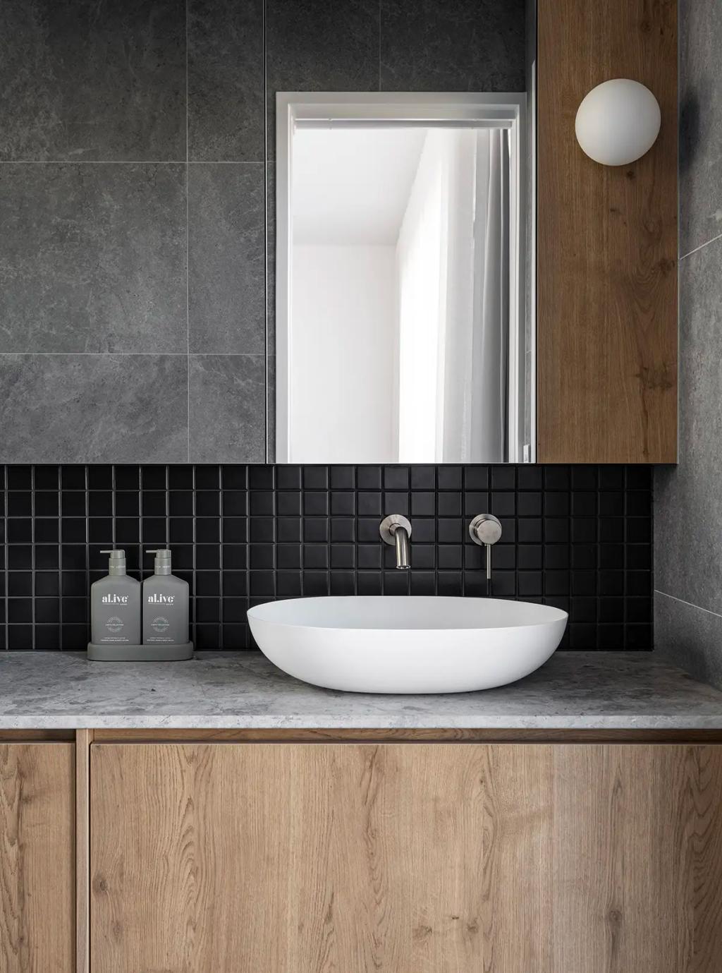 Project Ward 2.0 (A modern bathroom with gray stone walls, a wood vanity, a round white vessel sink, and black square tiles)