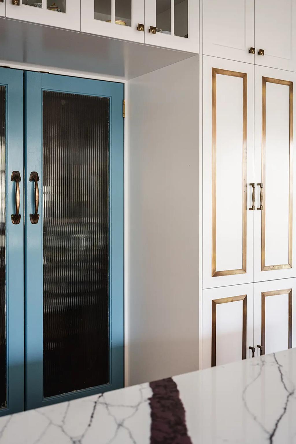 Project Coral Sea (White kitchen cabinets with brass accents, blue glass-paneled doors, and marble countertop in foreground)