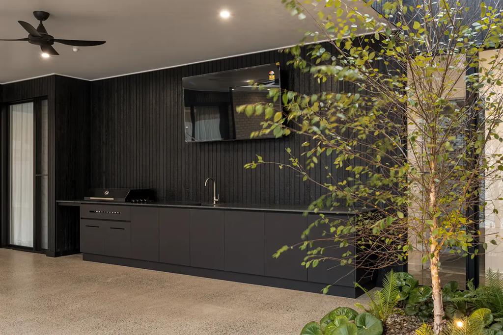 Modern black outdoor kitchen with TV and greenery.