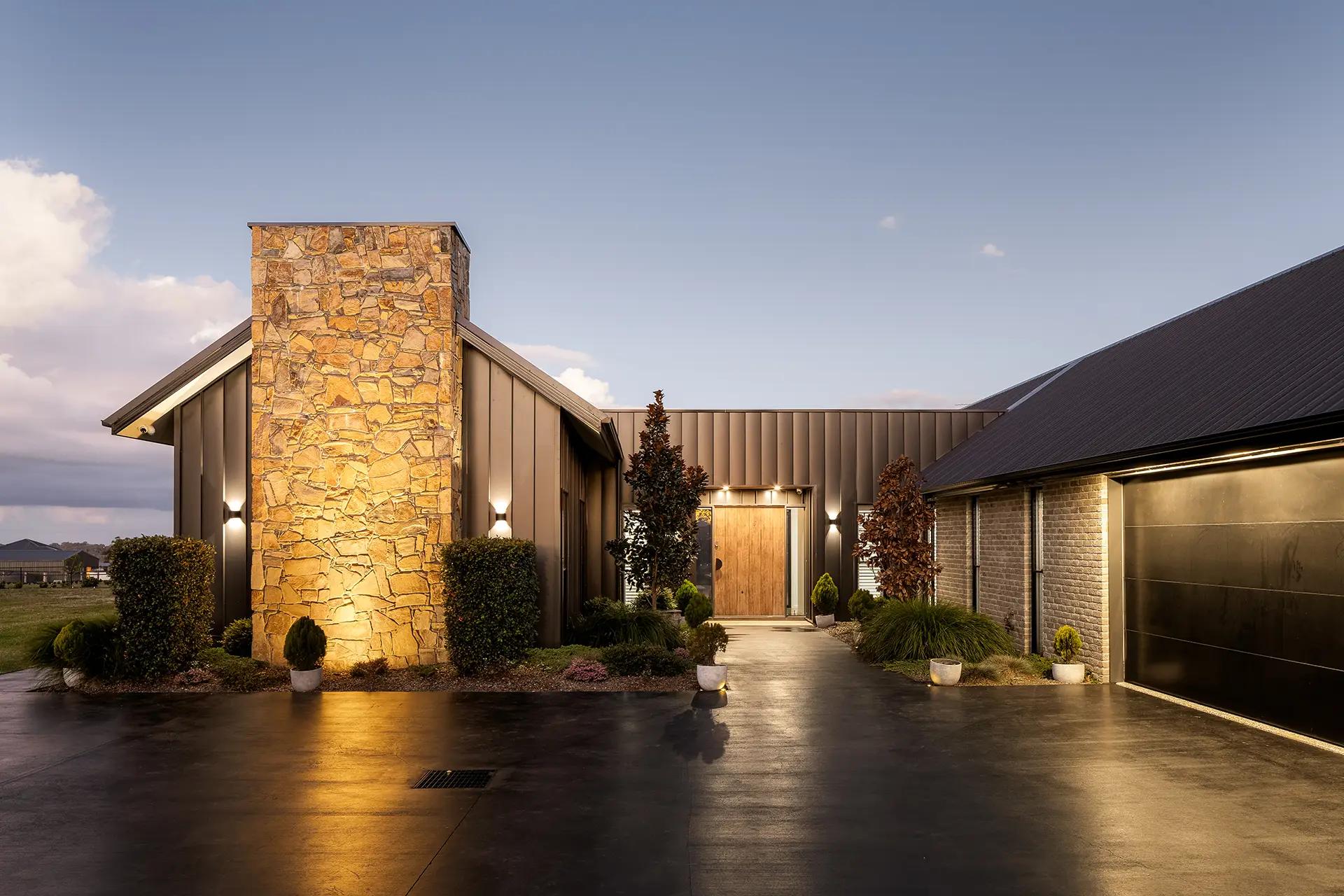 Project Mount Rascal (Modern home exterior with stacked-stone chimney, dark cladding and timber front door at dusk)