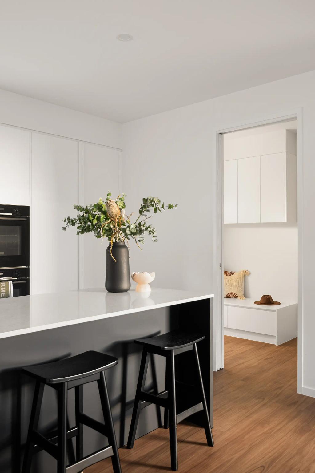 Project Ward (A modern kitchen with black stools, a white countertop, and a vase with green foliage, leading to a cozy room)