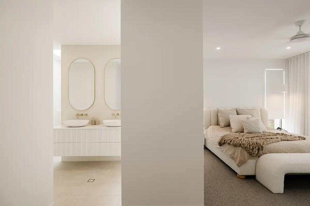 Project Vuse (A modern bedroom with a neutral color palette and a bed with beige linens and a chunky knit throw, adjacent to an ensuite bathroom with twin oval mirrors and white vanity units)