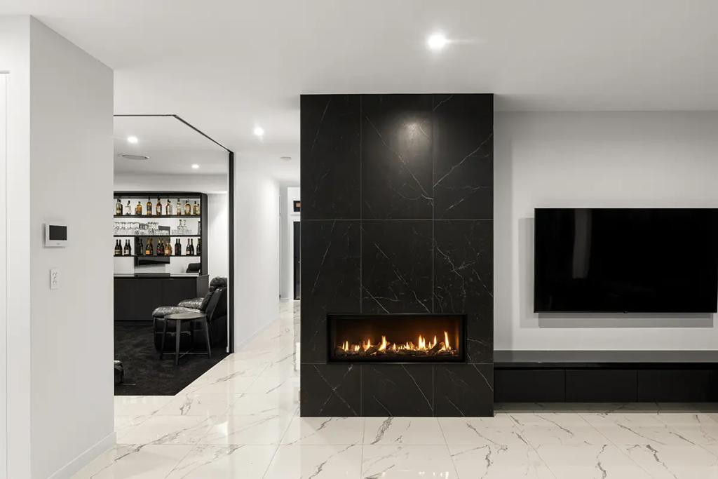 A modern living room with a black marble fireplace, flat screen TV mounted on the wall, and a home bar with bottles and glasses on the shelves in the background