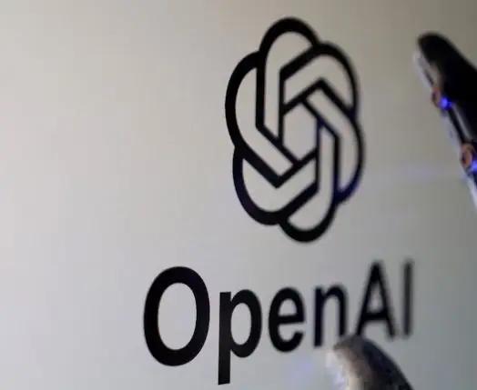 A close-up shot of a computer screen displaying the black OpenAI logo and text. A robotic mechanical finger with subtle blue glowing lights is touching the edge of the screen, symbolizing the intersection of robotics and artificial intelligence