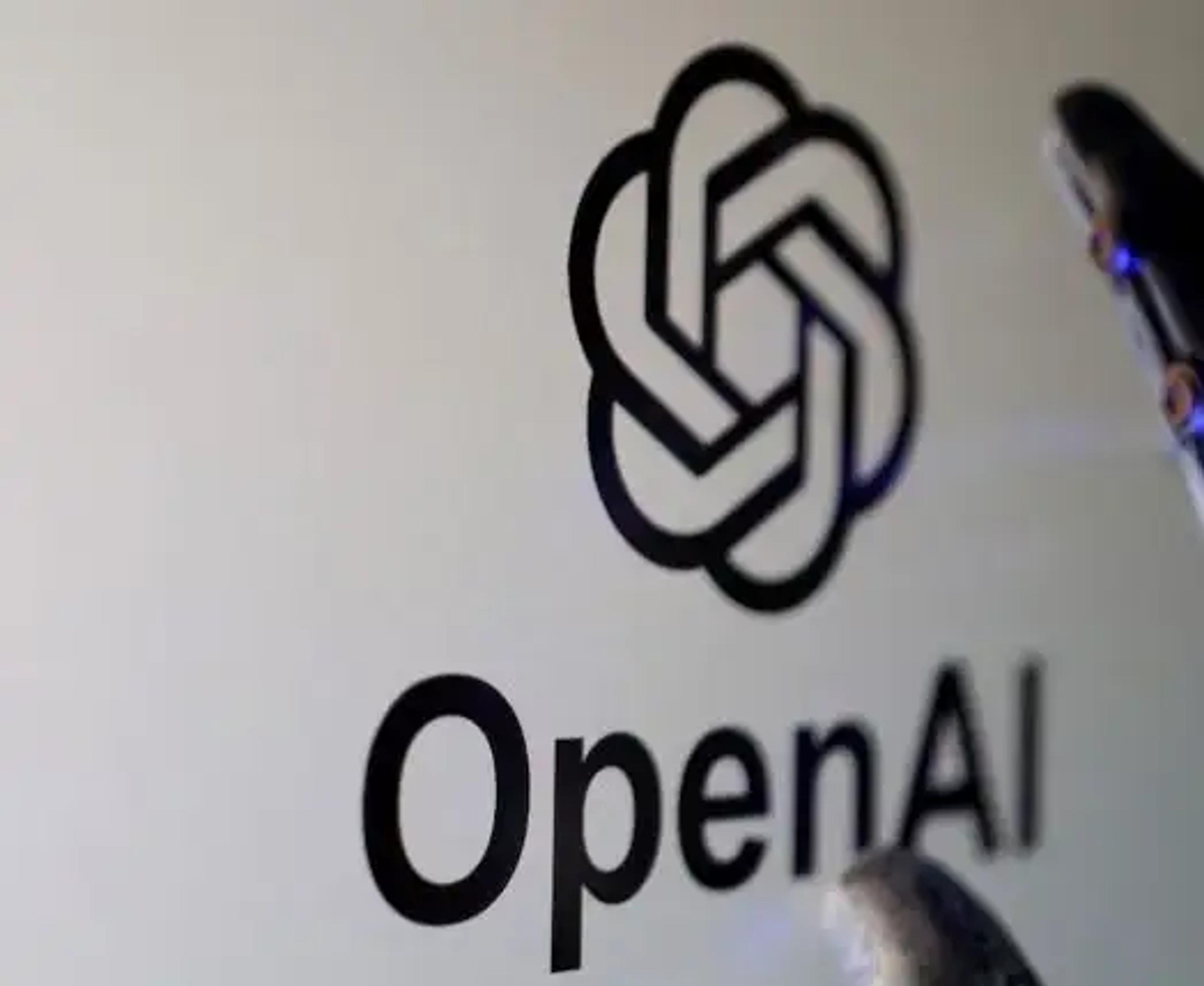 A close-up shot of a computer screen displaying the black OpenAI logo and text. A robotic mechanical finger with subtle blue glowing lights is touching the edge of the screen, symbolizing the intersection of robotics and artificial intelligence