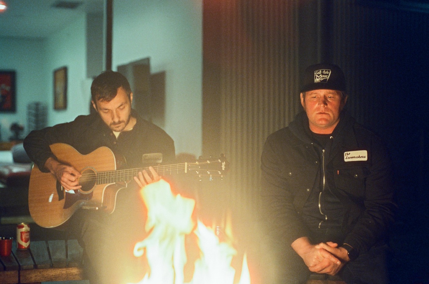 two men are sitting around a campfire playing guitar.