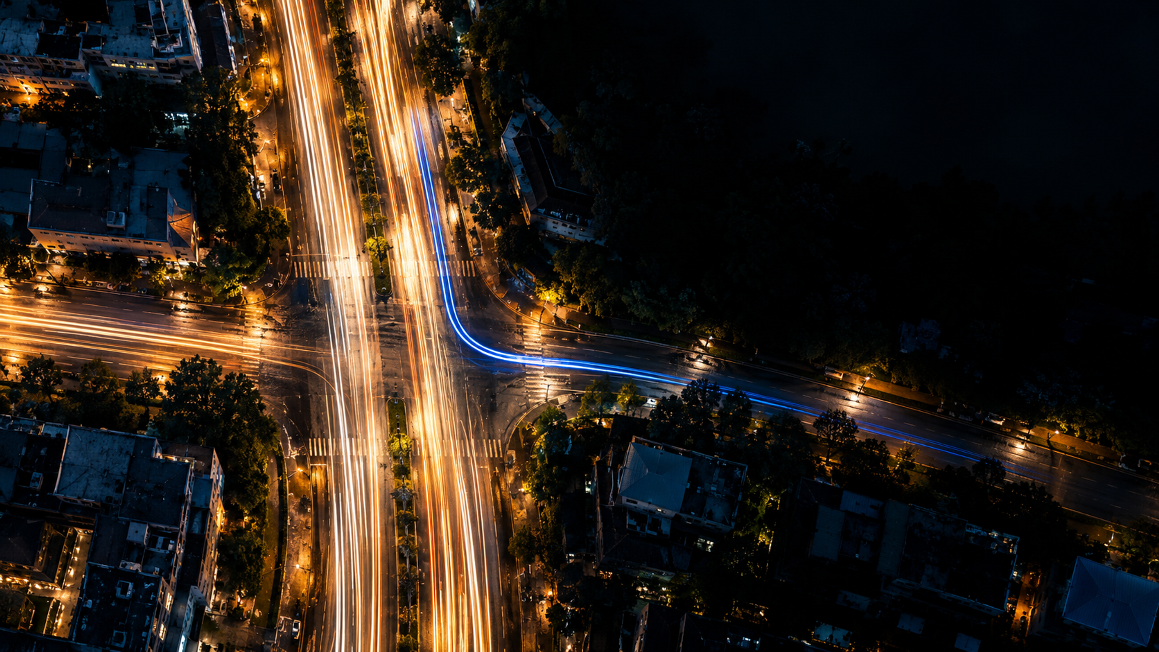 Aerial night view of a city intersection: amber light trails run straight through, and one blue trail turns off into a side street.