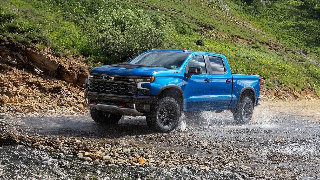 Blue Chevrolet Silverado 1500 driving through a shallow stream in a mountain landscape.