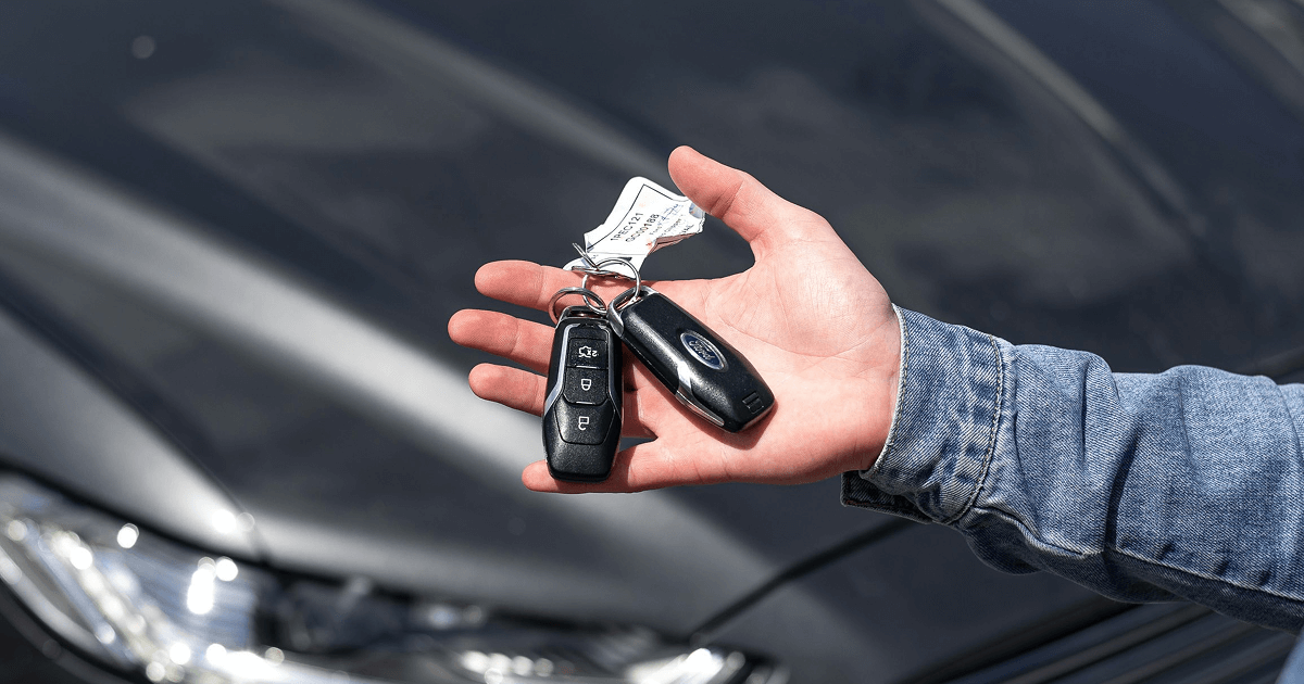 Hand holding a set of car keys in front of a parked vehicle