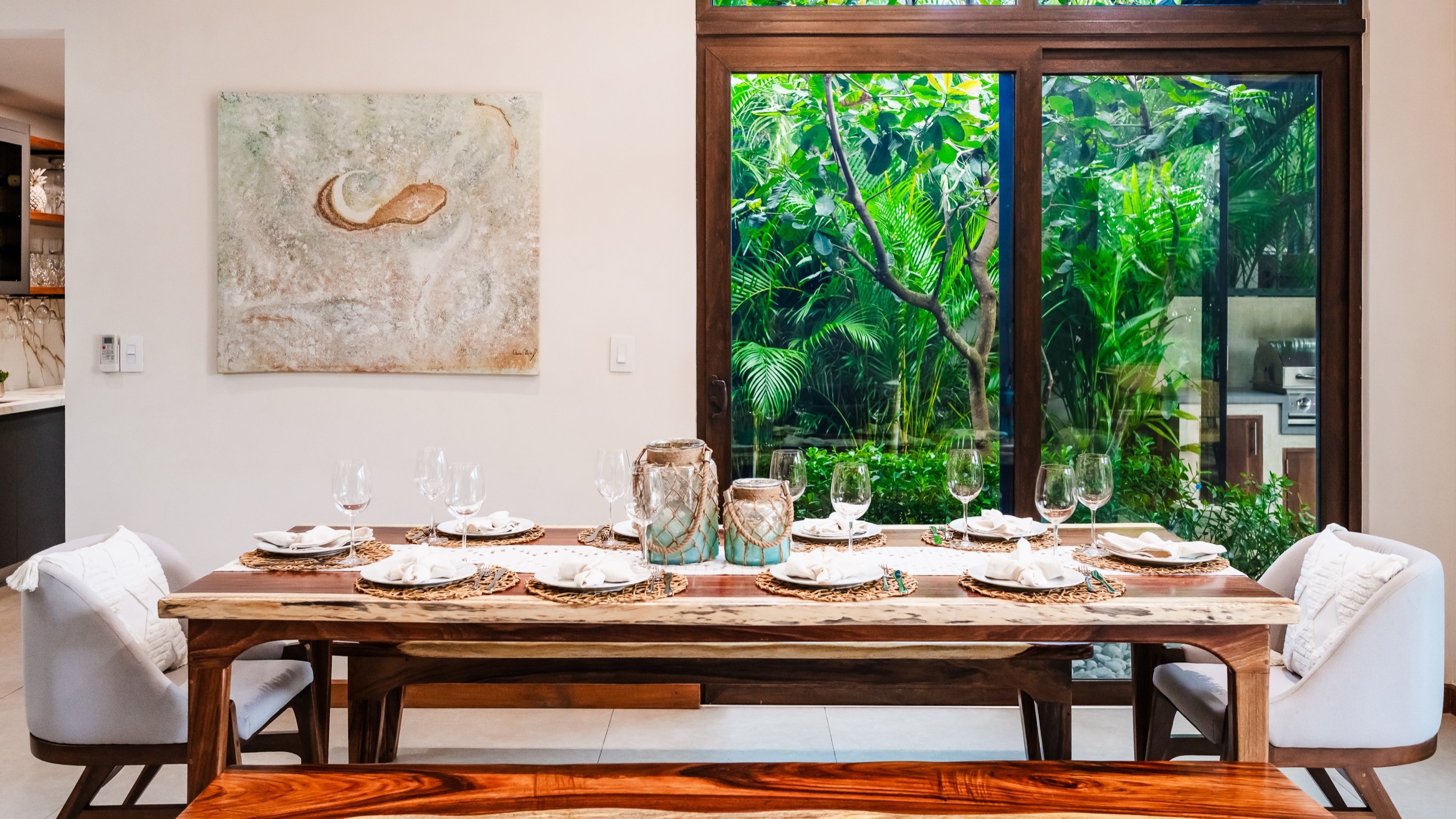 Dining table set for eight facing a floor-to-ceiling window onto tropical garden