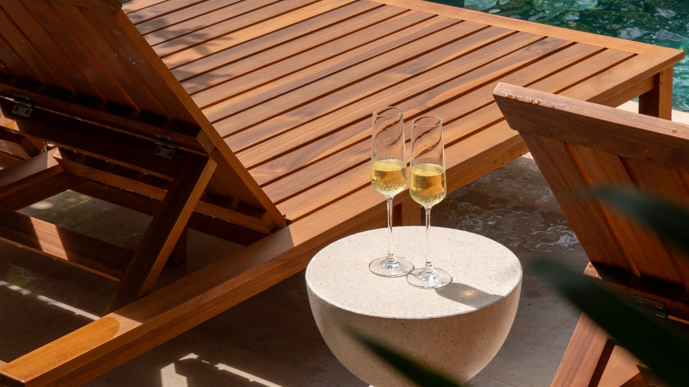 Two glasses of sparkling wine on a side table beside a teak sun lounger and pool