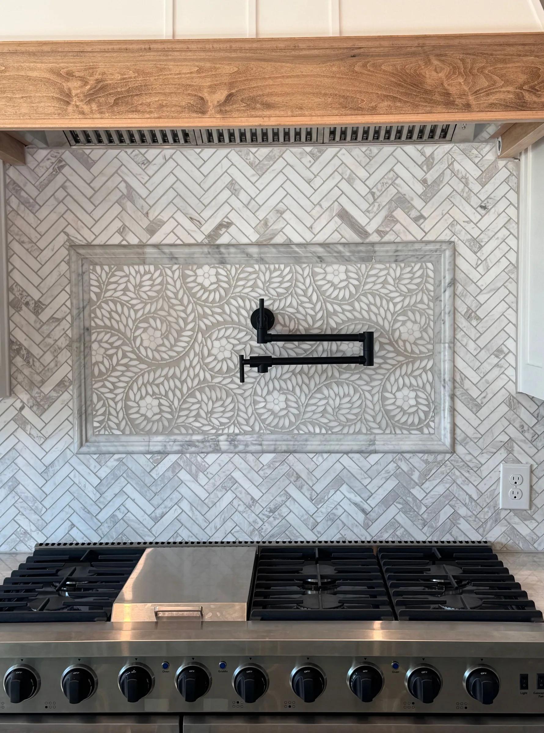 Marble herringbone backsplash with a framed mosaic panel and a pot filler over a gas range