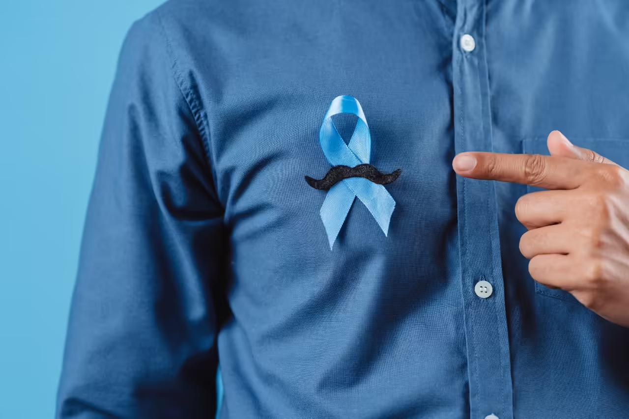 Close-up of a blue awareness ribbon with a small black moustache pinned to a man’s dark blue shirt, as he points towards it