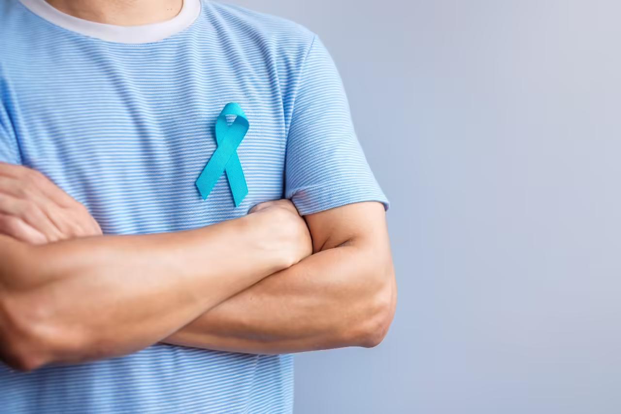 A man in a pale blue striped T-shirt stands with his arms folded, wearing a blue prostate cancer awareness ribbon