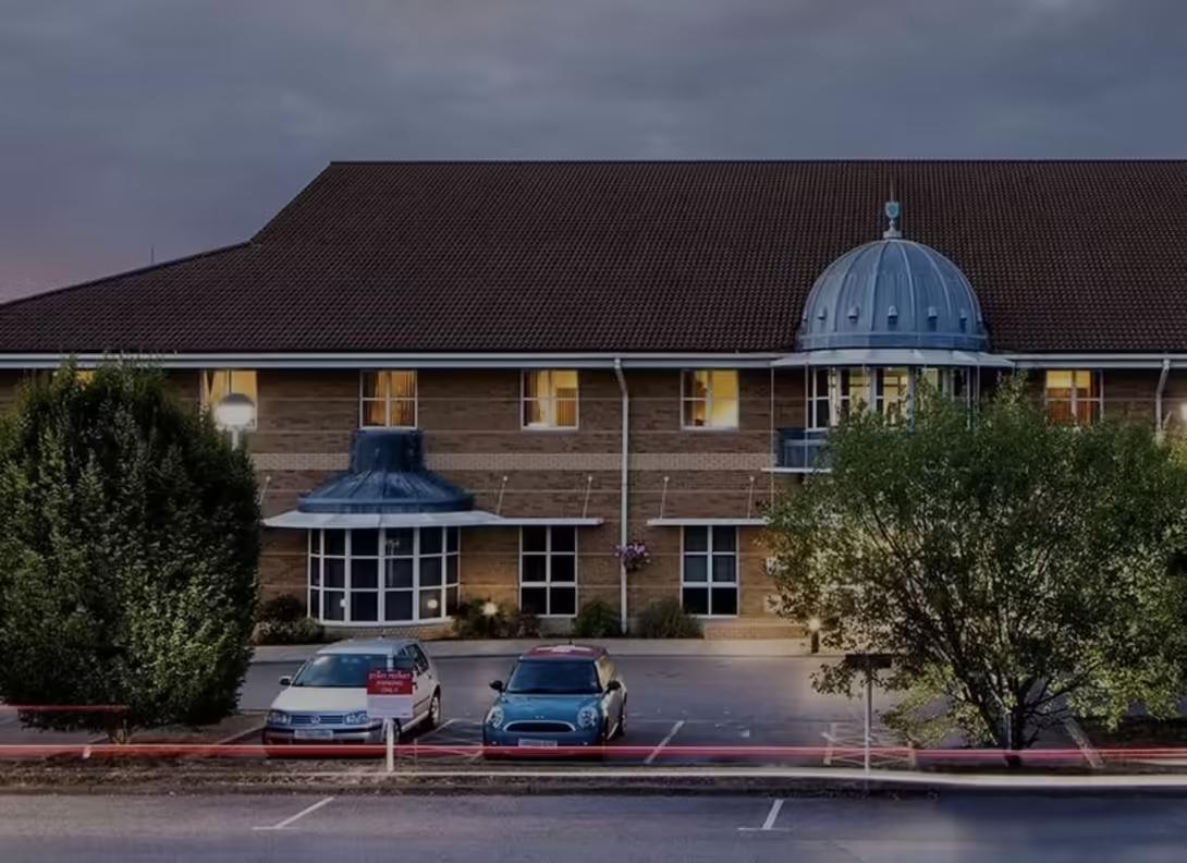 The two-storey stone frontage of Nuffield Health Guildford at dusk, with its domed entrance rotunda and cars in the car park