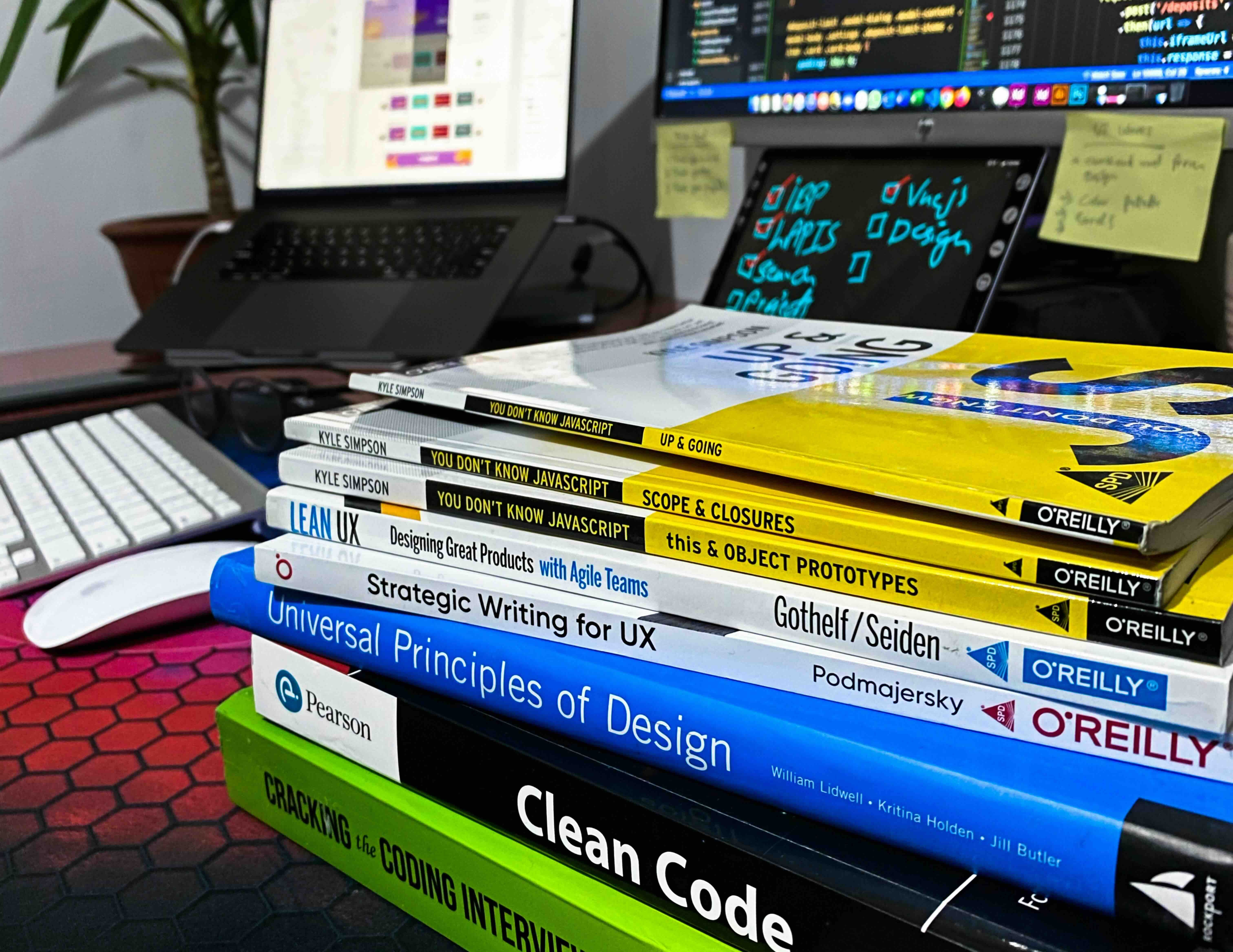 A stack of software engineering and design books on a desk in front of monitors showing code, including Clean Code and Cracking the Coding Interview.