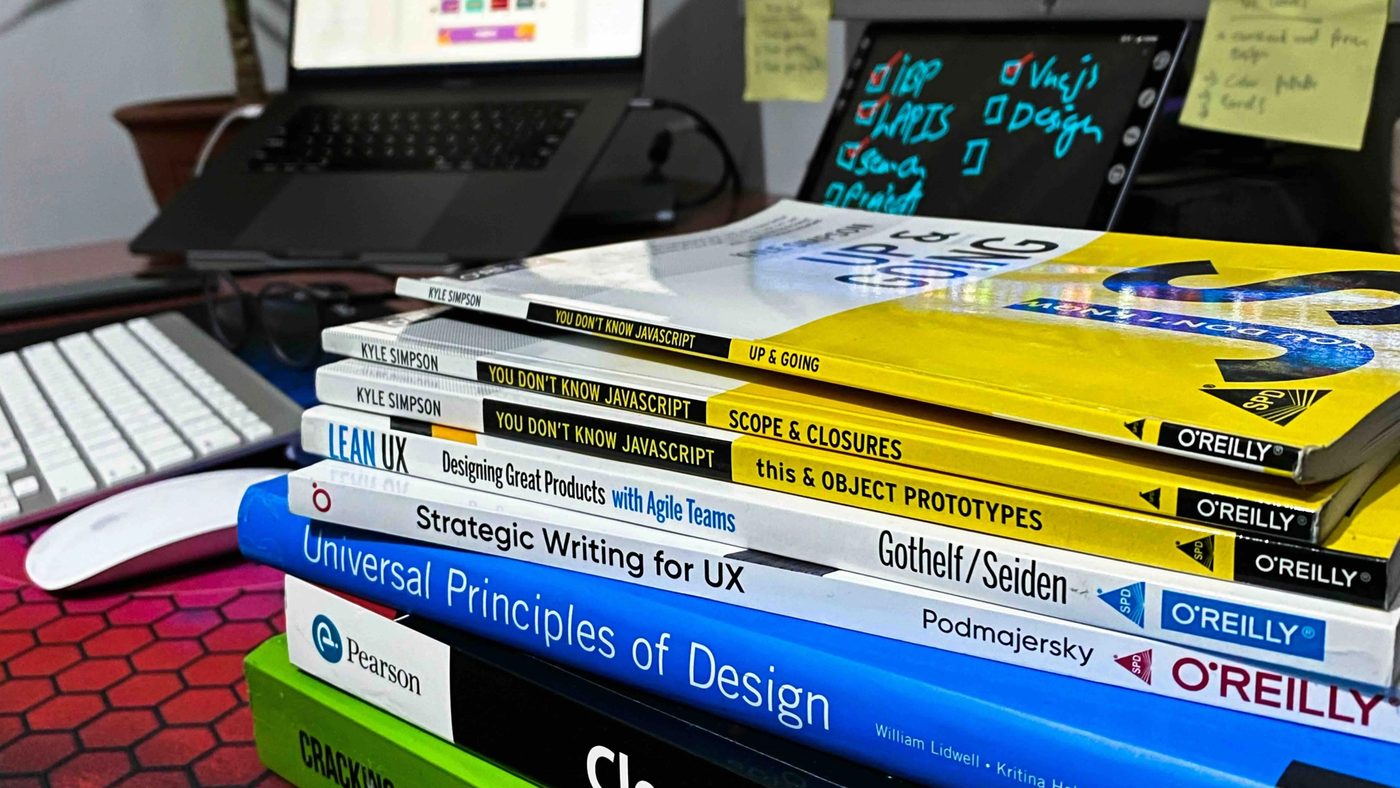 A stack of software engineering and design books on a desk in front of monitors showing code, including Clean Code and Cracking the Coding Interview.