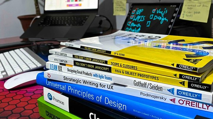 A stack of software engineering and design books on a desk in front of monitors showing code, including Clean Code and Cracking the Coding Interview.